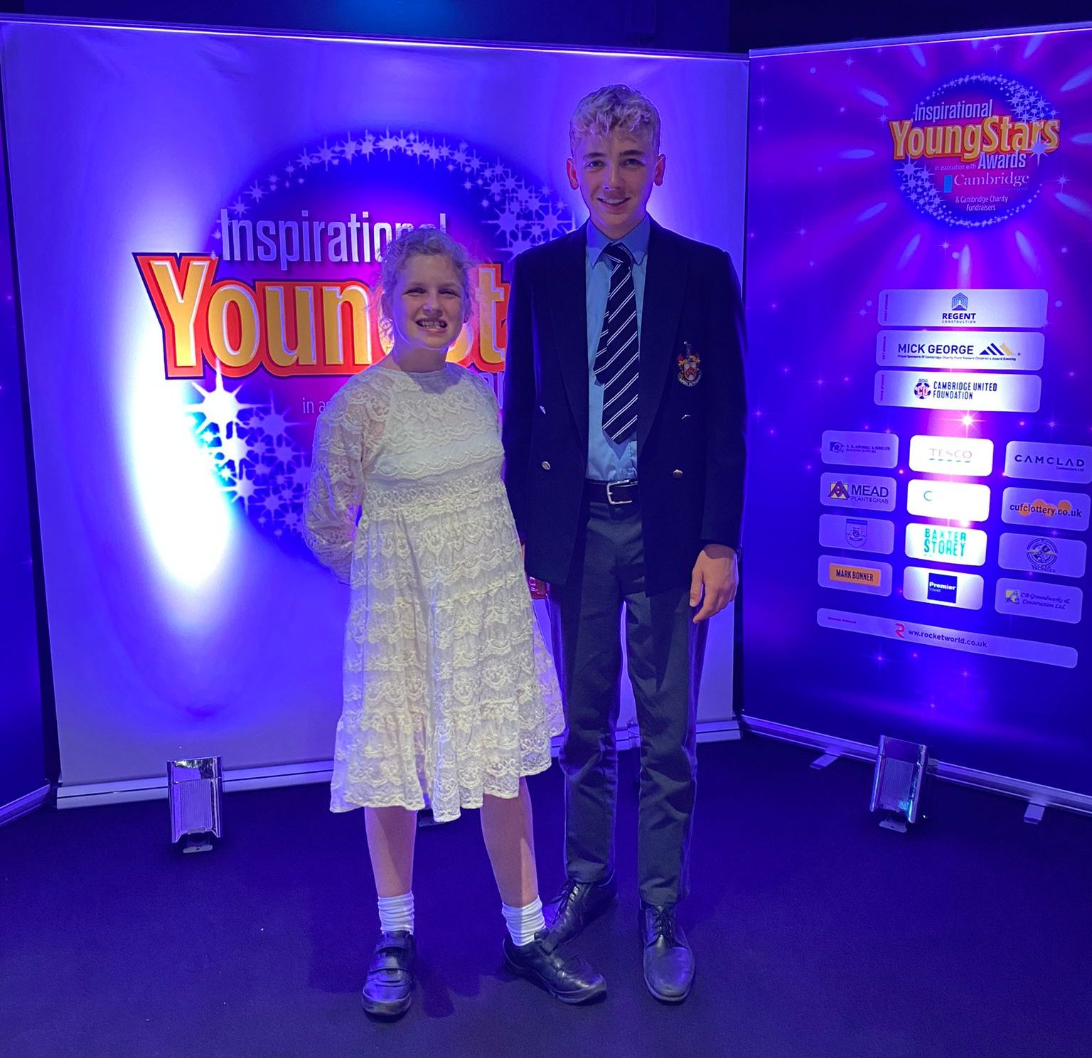 Young Ambassadors Mimi and Flint Clarke winning an award for their support of Tom's Trust. Flint is wearing a suit and tie and Mimi is wearing a white dress and they are on a stage with blue lighting in the background.